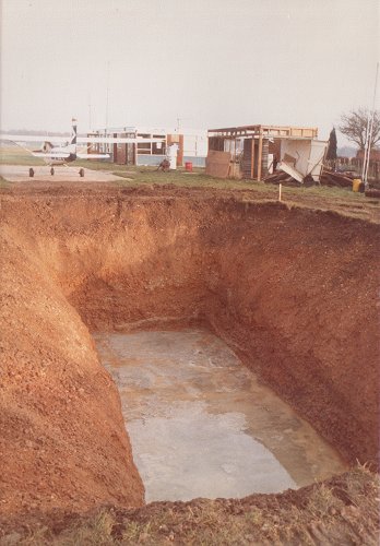 The excavation for Tank 7, is shown with its concrete base lining. The partially demolished fire station building can be seen on the right. Tank 1 and Tank 4 are underground near the pump.